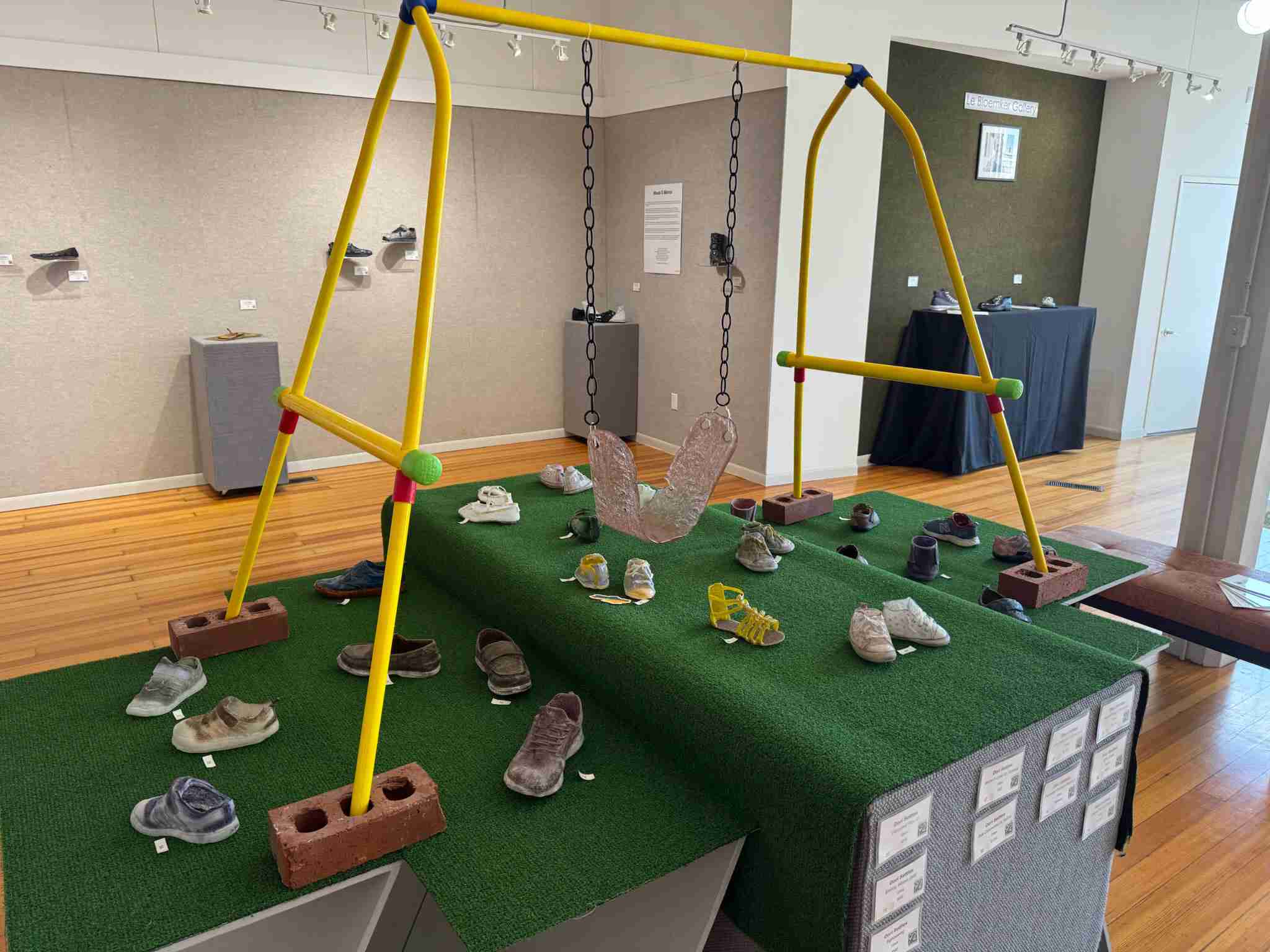 Glass children's shoes staged under a yellow model swingset with a glass swing, in an art gallery