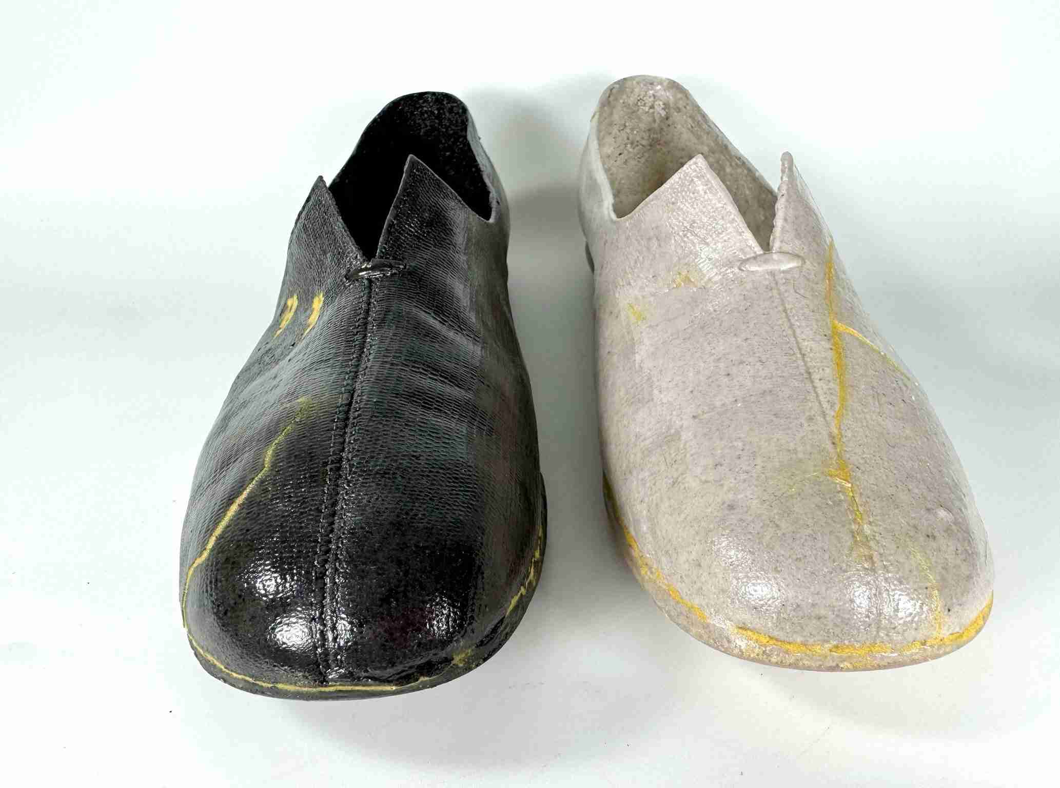 Black glass shoe with gold-filled cracks on the left and a pale peach colored glass shoe with gold-filled cracks on the right, facing forward.
