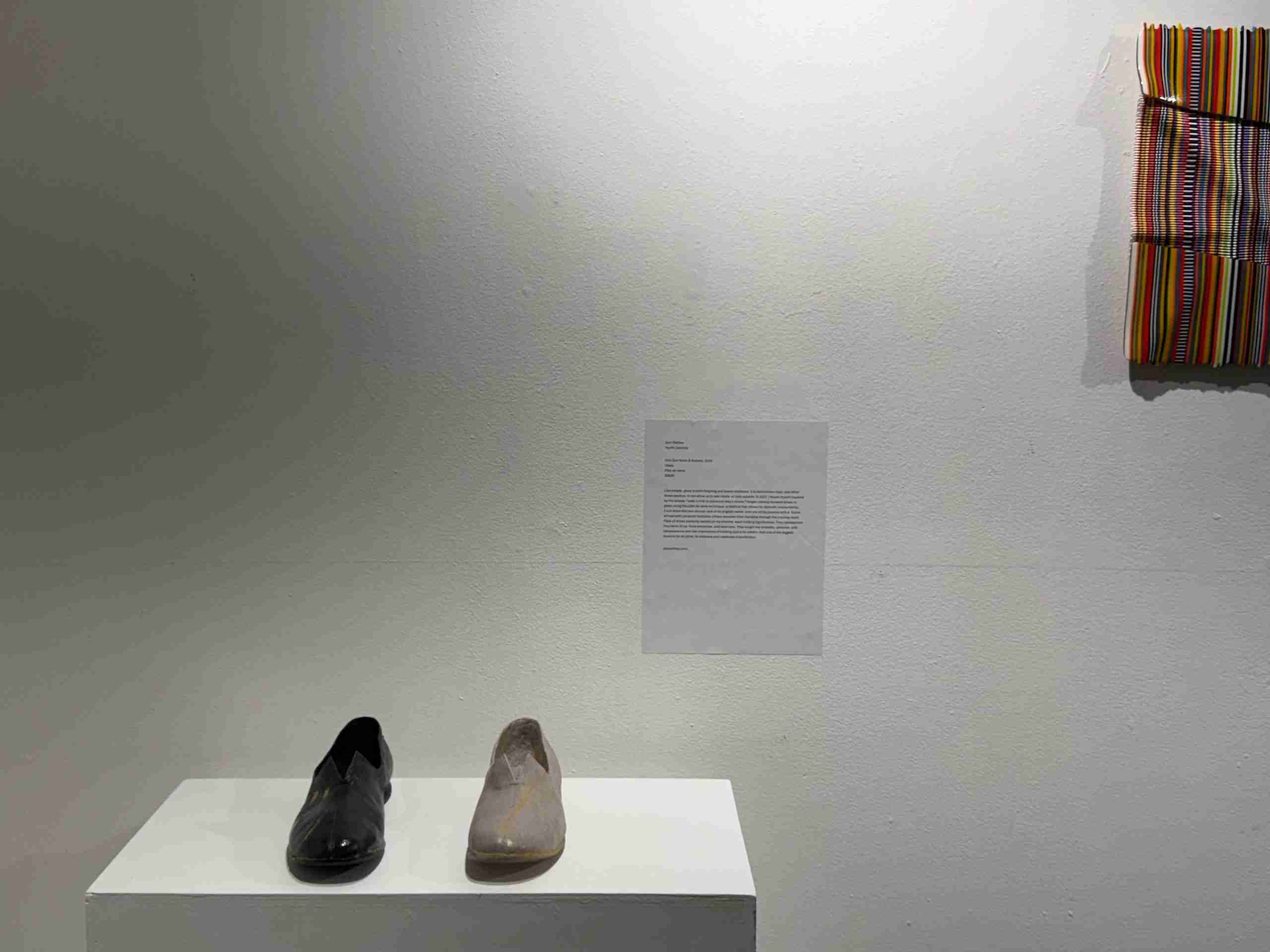 Pair of shoes, one black one beige on a pedestal with an artist statement behind it and another piece of artwork to the right