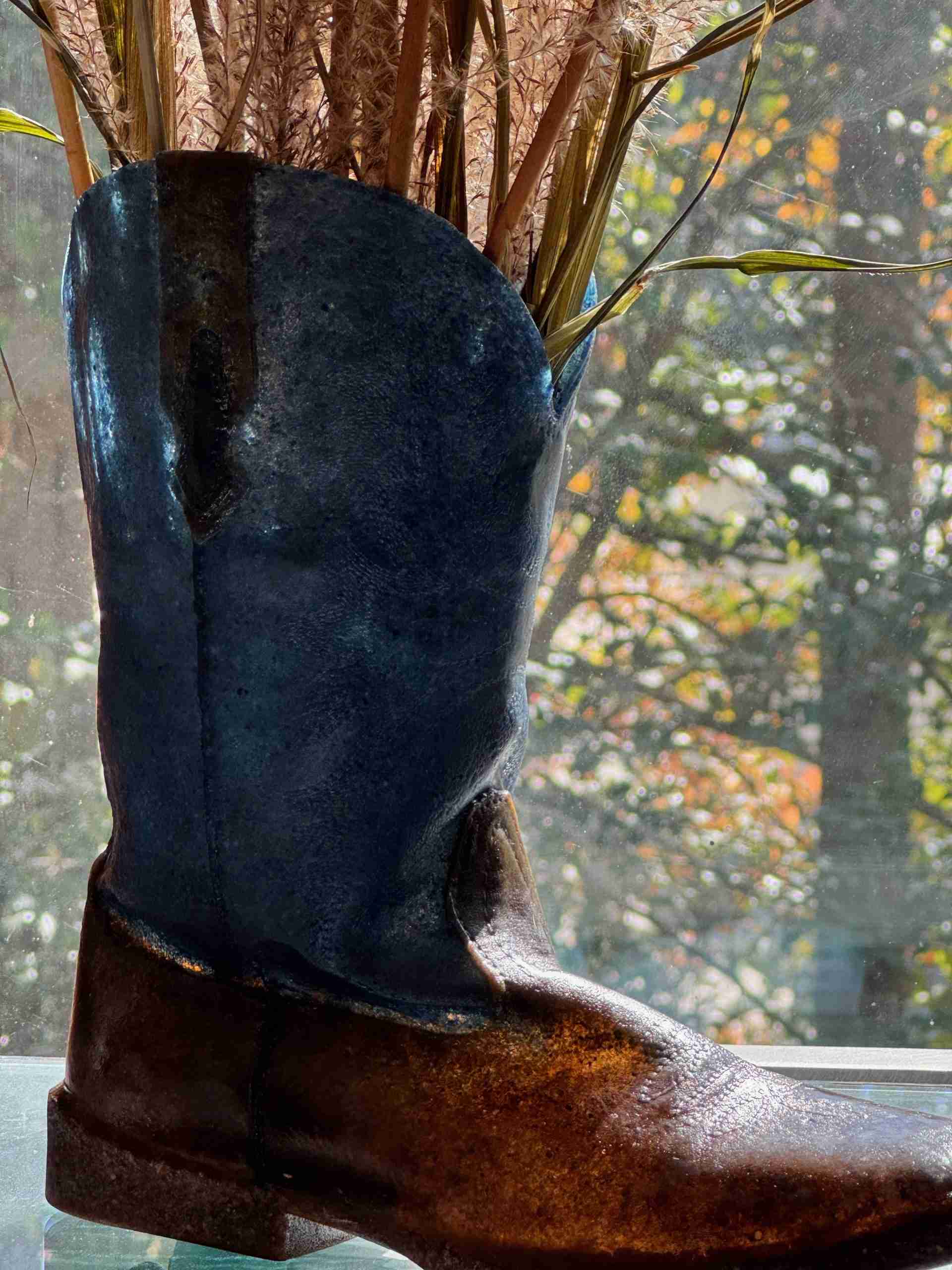 Glass Blue and Brown Cowboy boot sitting in front of a window with dried grasses set inside like a vase created by Dori Settles